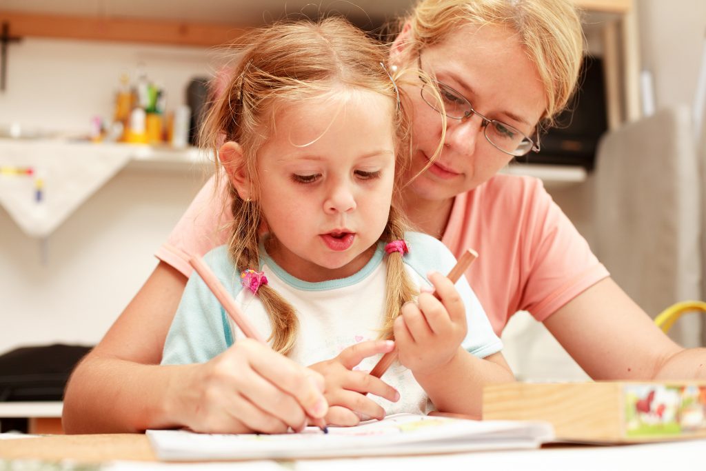 A mother sits beside her young daughter, teaching the girl to write with a pencil.