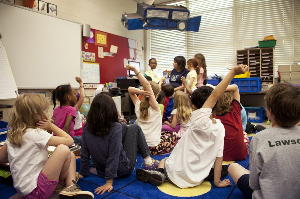 School children seated on the floor before a teacher at the front of the room.